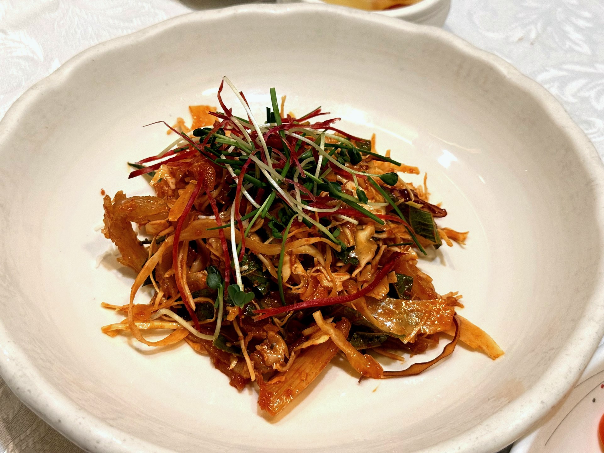 Sudam Hanjeongsik 무침 (seasoned cold dish) - shredded vegetables and seafood tossed in a glossy mild red chili-and-sesame dressing, topped with fine red and green sprouts, on a white ceramic bowl