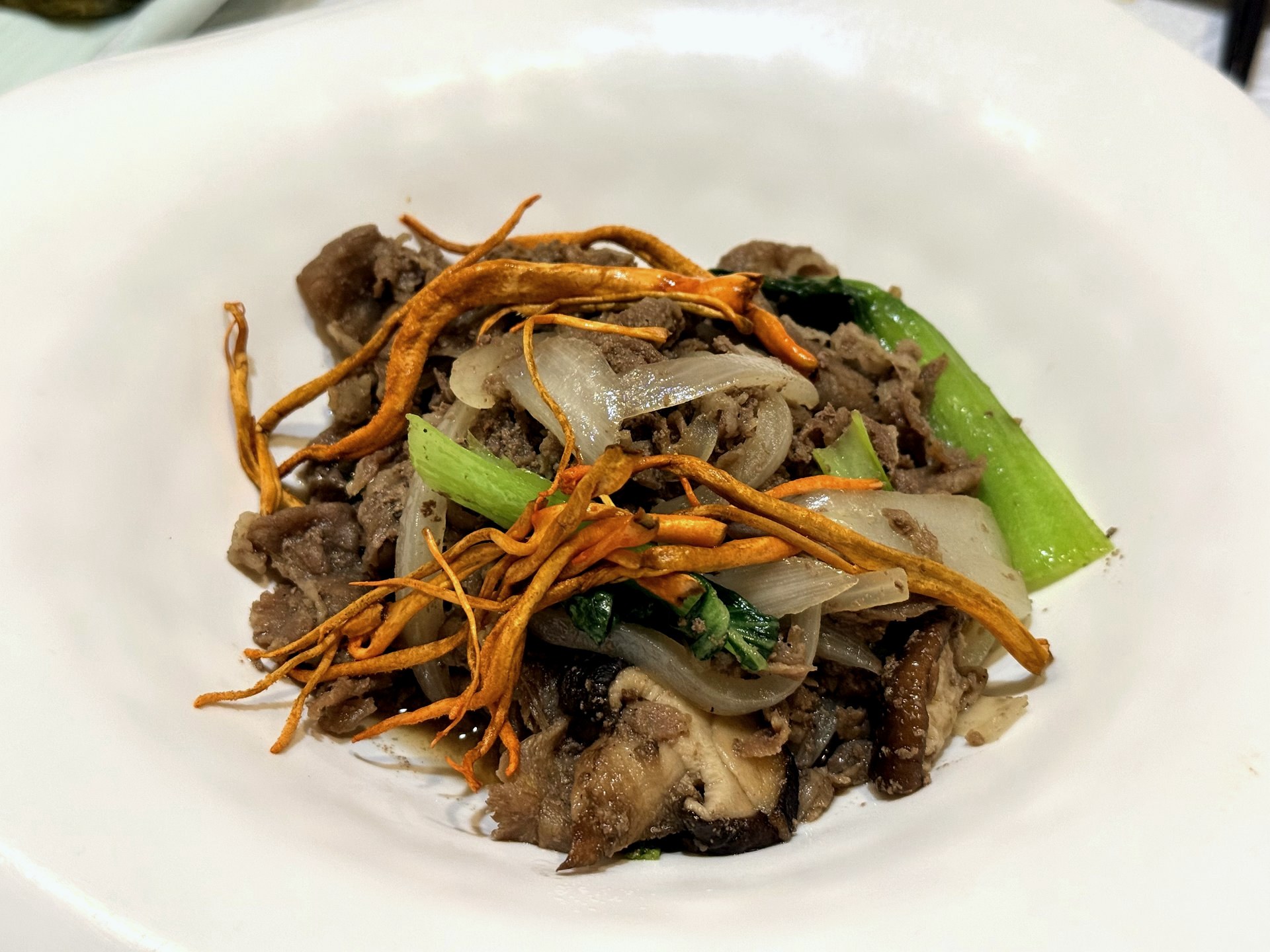 Sudam Hanjeongsik 불고기 (bulgogi) - thin slices of marinated beef stir-fried with sliced onion, shiitake mushroom and bok choy, topped with crisp golden cordyceps mushroom strands, on a white plate