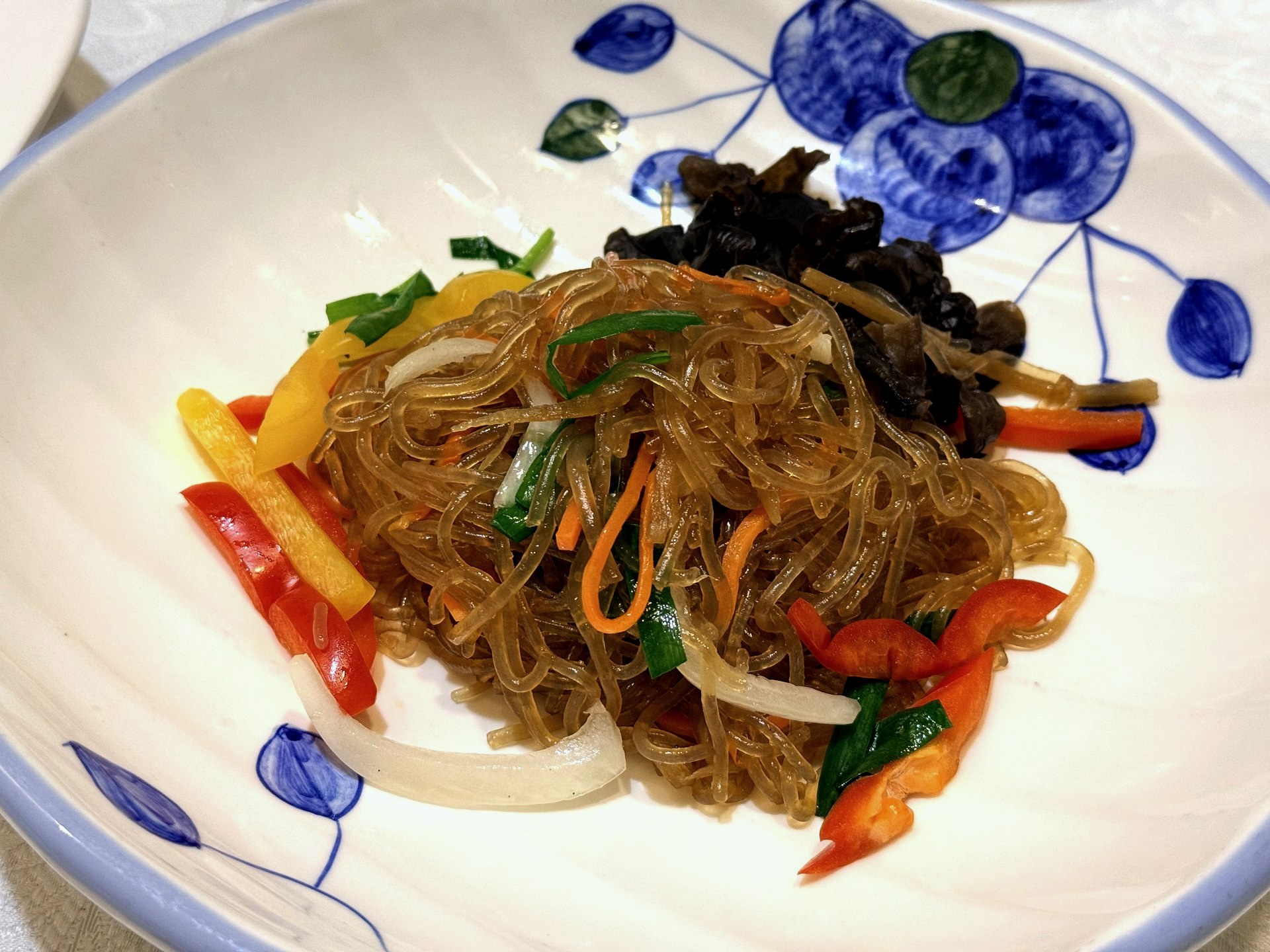 Sudam Hanjeongsik 잡채 (japchae) - glossy stir-fried sweet-potato glass noodles with red and yellow pepper strips, onion, chive and dark wood-ear mushroom, on a blue-and-white floral bowl