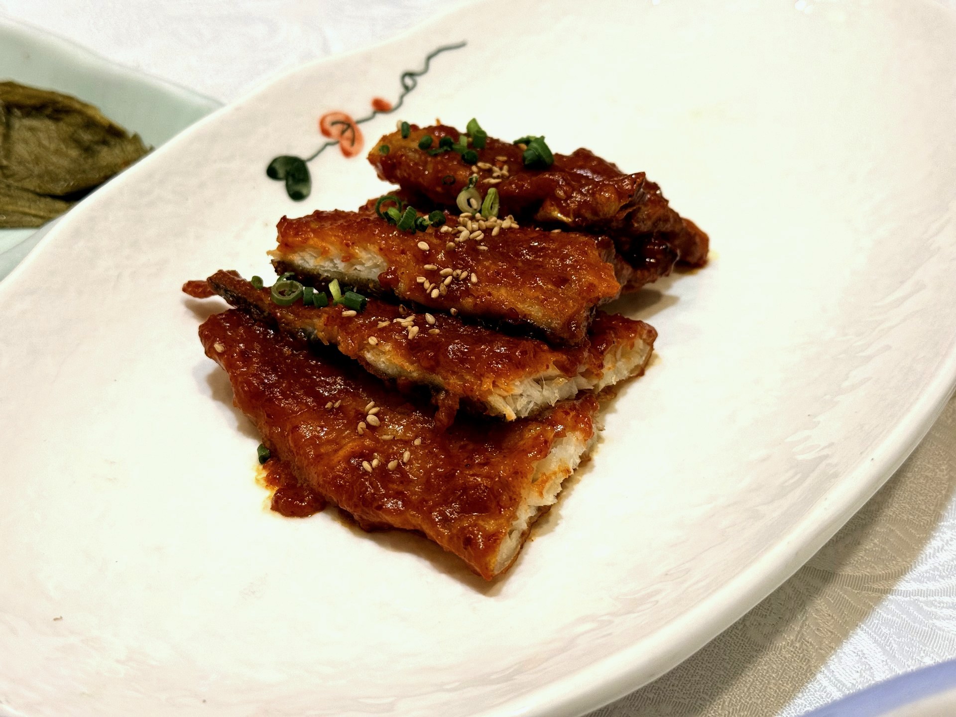 Sudam Hanjeongsik 코다리 (half-dried pollock) - meaty fillets of pollock glazed in a glossy sweet-spicy red gochujang sauce, garnished with chopped scallion and sesame seeds, on a floral-rimmed white plate