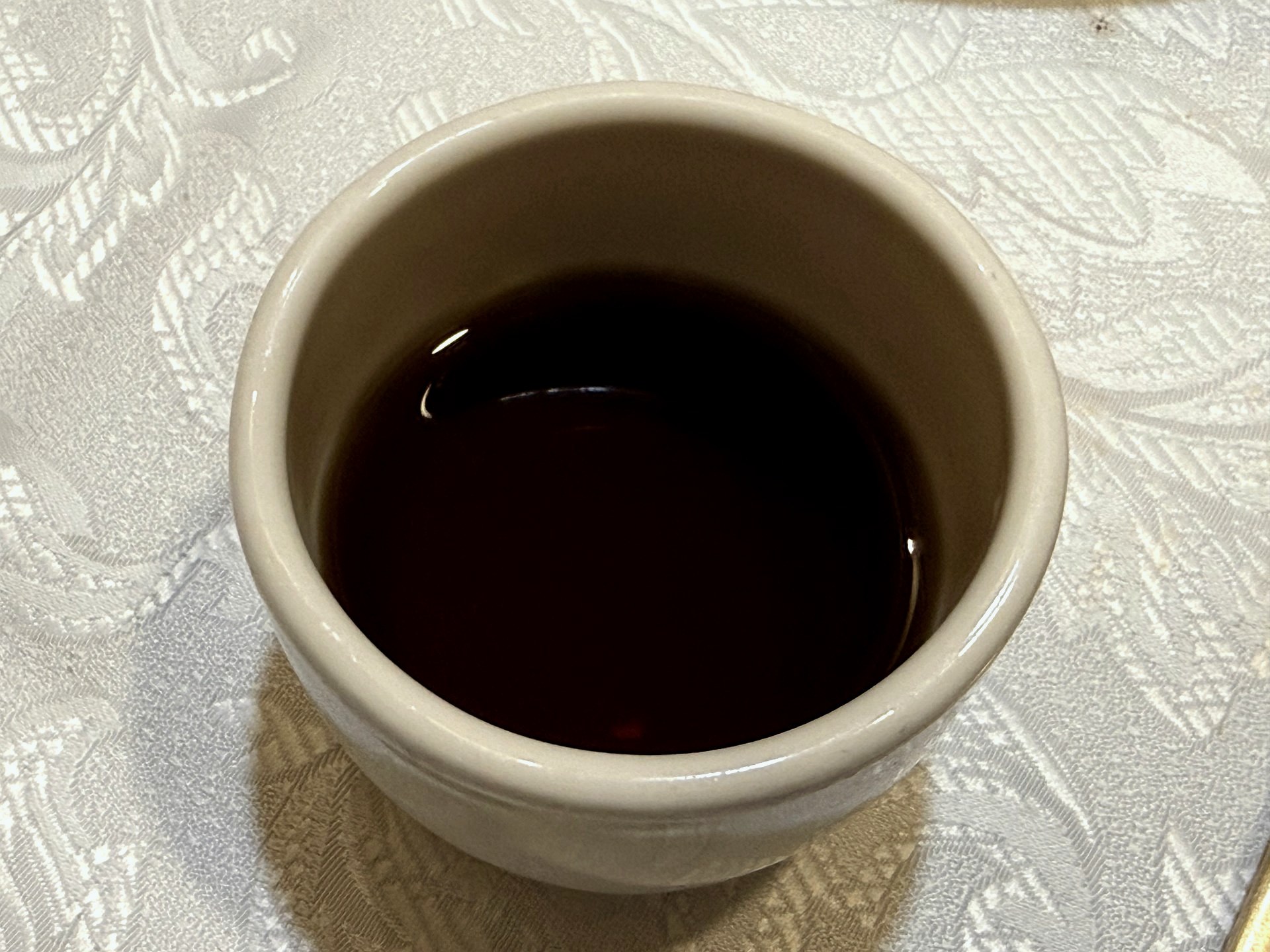 Sudam Hanjeongsik 매실차 (green-plum tea) - a small white ceramic cup of clear dark-amber plum tea on a white damask tablecloth, served to finish the meal