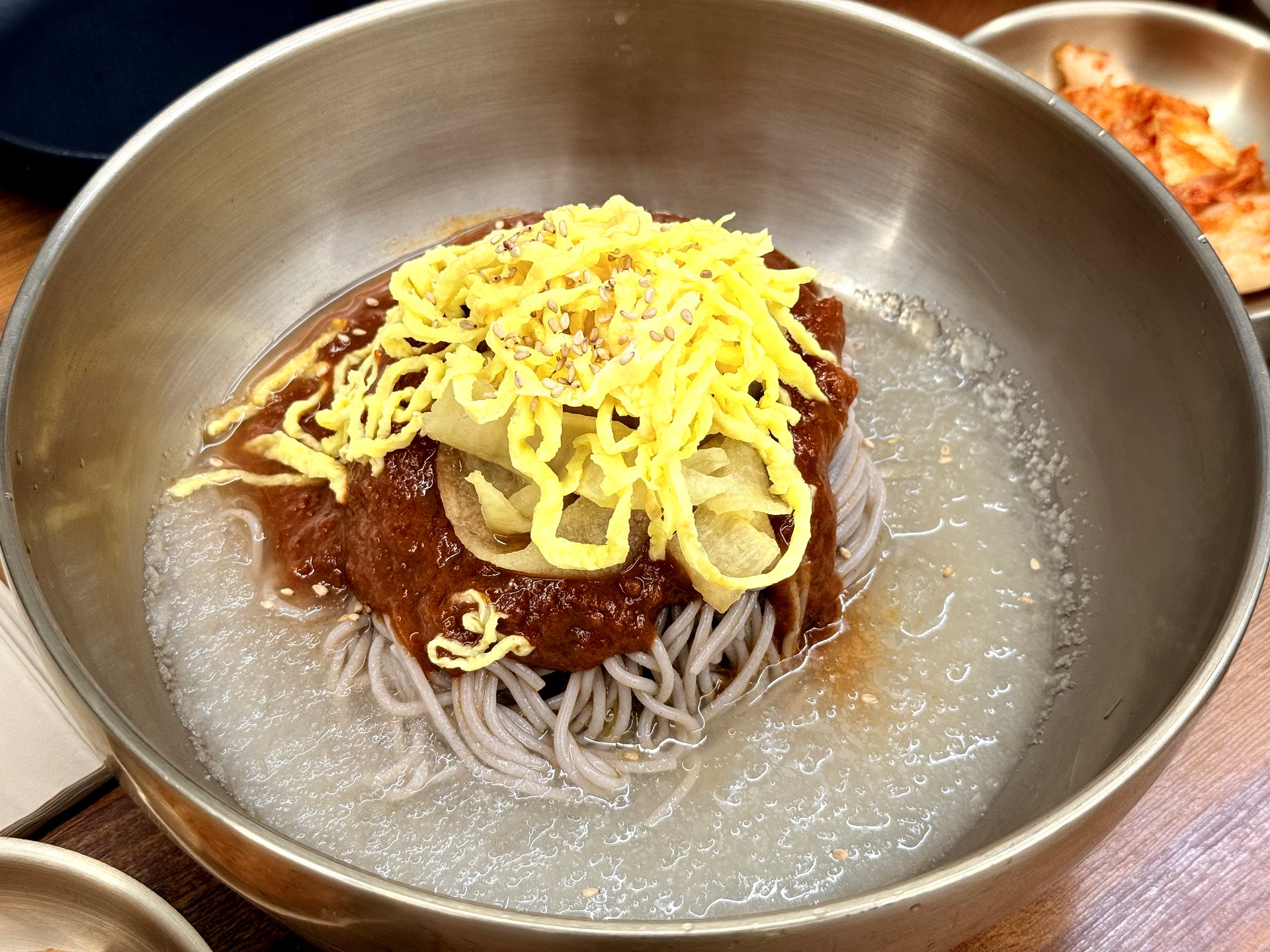 Memilbat-e Gasiri bibim makguksu - buckwheat noodles in sweet-spicy red gochujang sauce topped with shredded egg and pickled radish, in a metal bowl