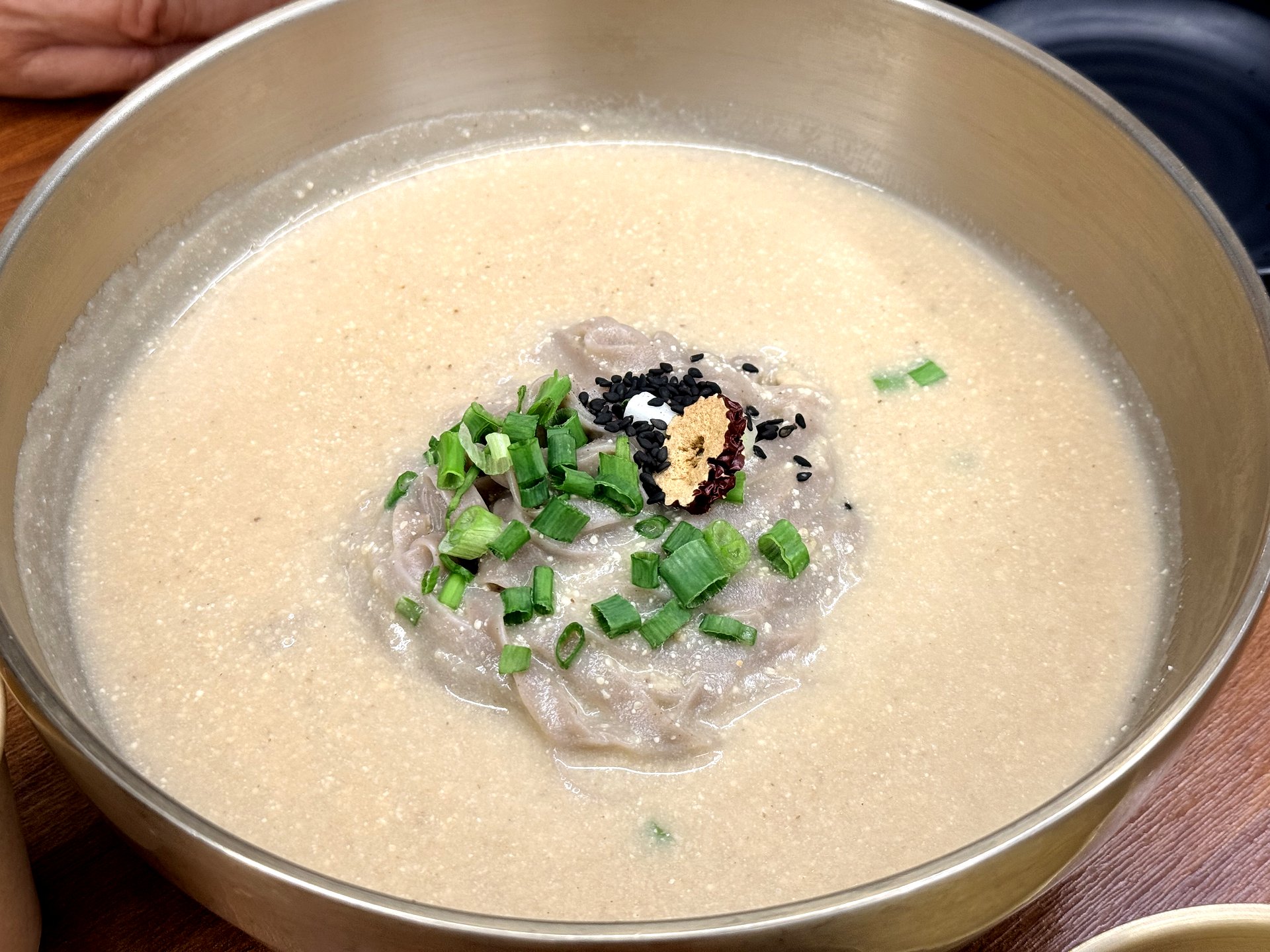 Memilbat-e Gasiri warm deulkkae makguksu - buckwheat noodles in a creamy pale ground-perilla-seed broth topped with scallion, jujube and black sesame, in a brass bowl