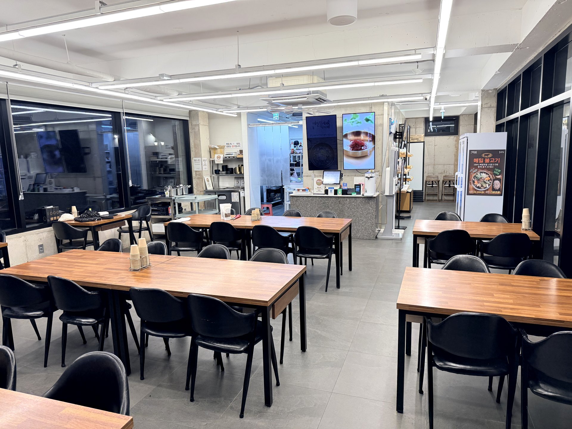 Bright industrial-style interior of Memilbat-e Gasiri Donggwang with wooden tables, an open kitchen and a buckwheat-bulgogi standee