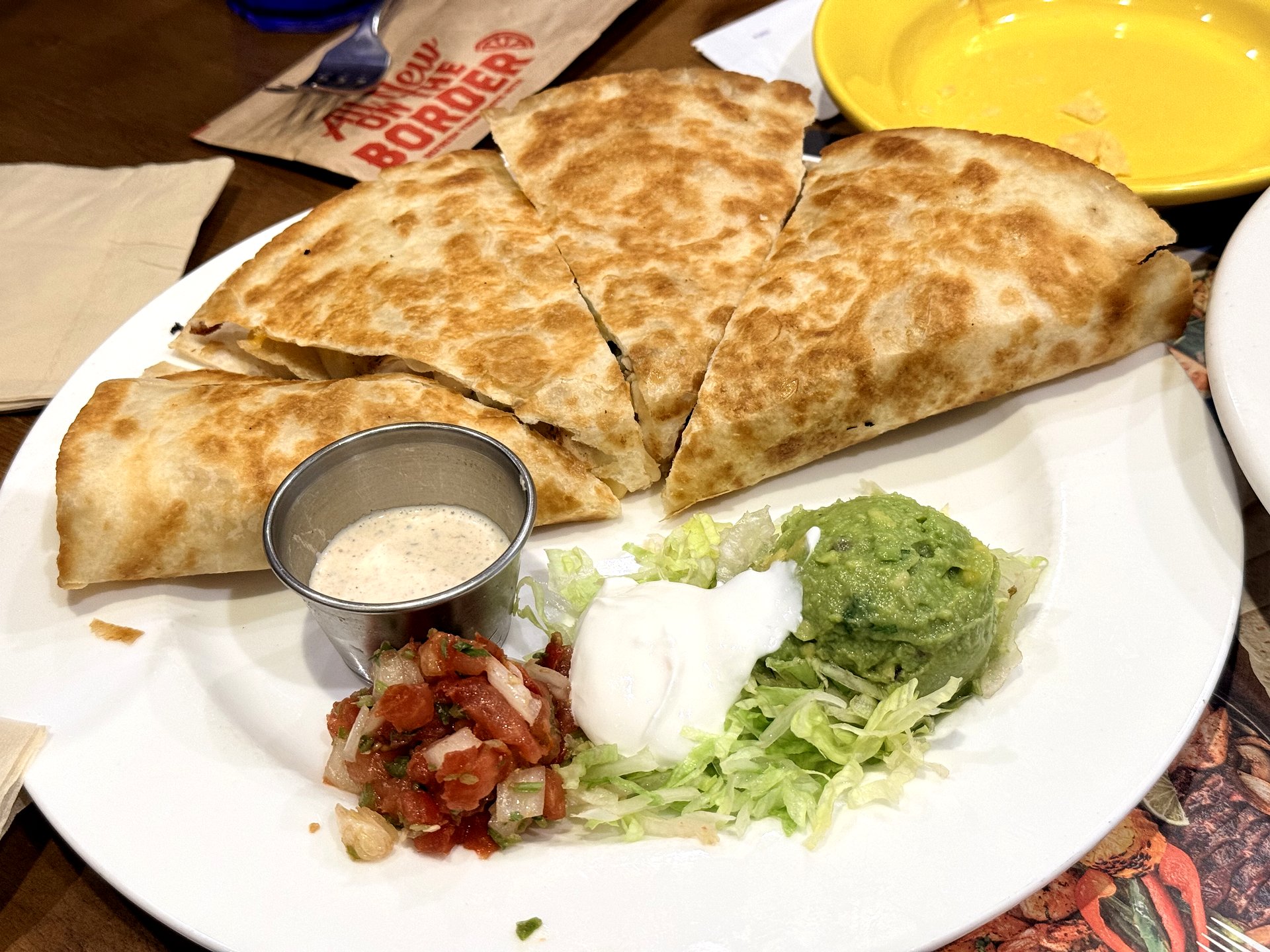 On The Border quesadilla plated - golden griddled flour-tortilla wedges on a white plate with a cup of ranch dip and a garnish of pico de gallo, sour cream, shredded lettuce and guacamole