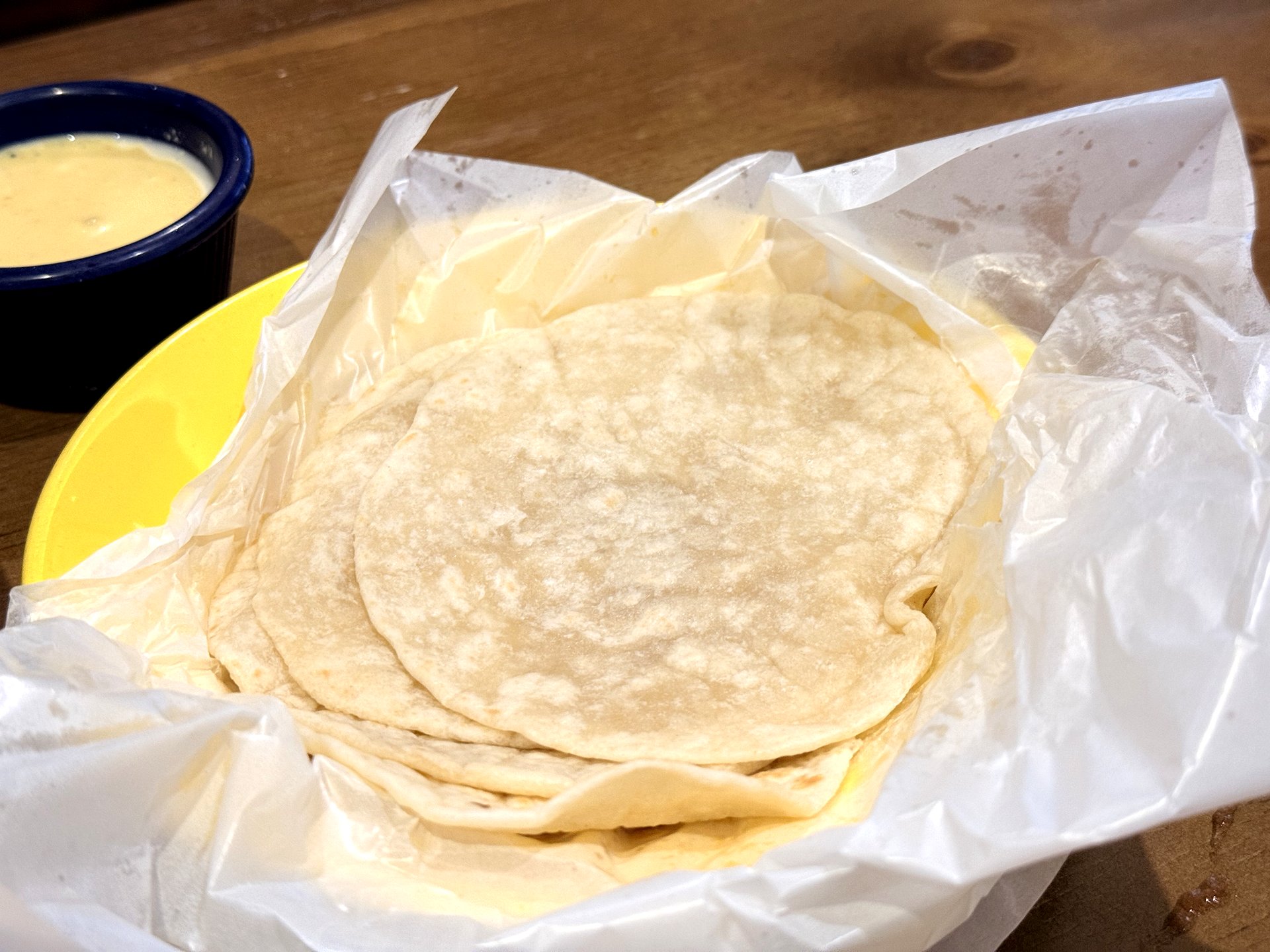 A basket of warm soft flour tortillas wrapped in paper next to a small dish of creamy queso cheese dip at On The Border, for building your own steak fajita wraps