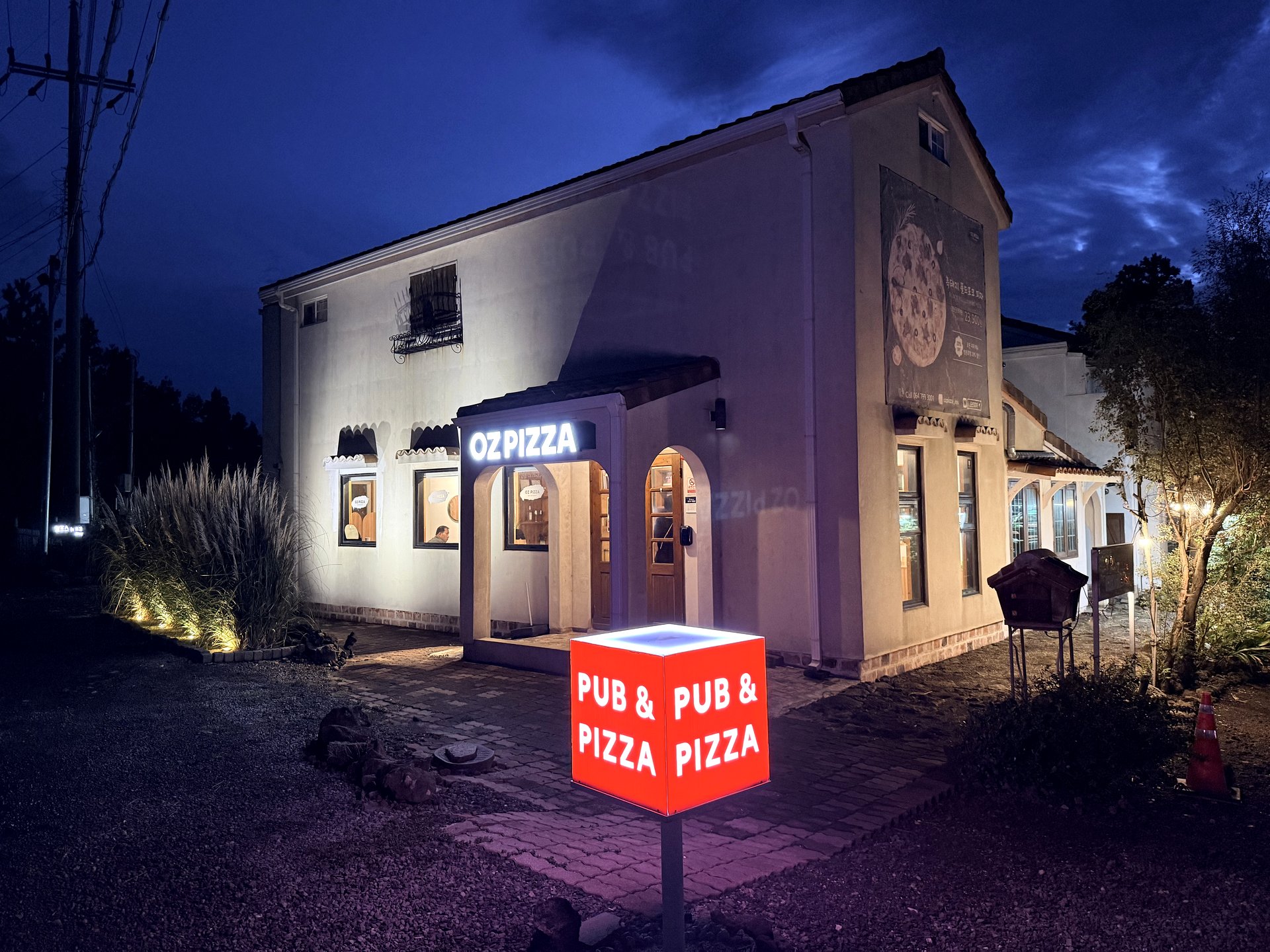 OZ Pizza exterior at dusk in Sogil-ri, Aewol, Jeju - a converted house with warm lit windows and a glowing red PUB and PIZZA sign out front