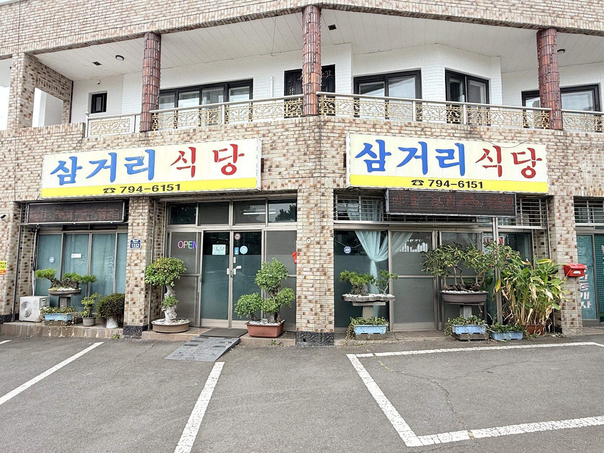 Samgeori Sikdang exterior in Andeok, Jeju - a corner restaurant with blue-and-red hand-painted 삼거리식당 signs, an OPEN sign, bonsai plants by the glass door, and a parking lot, at address Ilju-seo-ro 1332